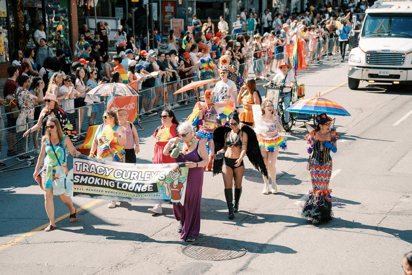 Participants in colorful costumes march in a lively parade, holding a banner for 'Tracy Curley's Smoking Lounge', as spectators watch from the sidelines.