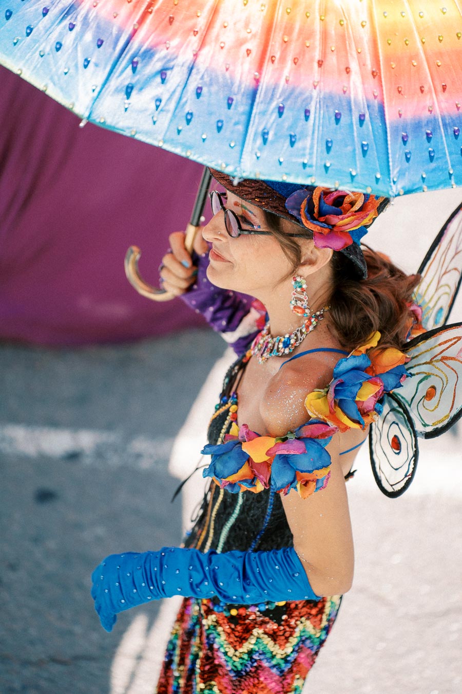 A person dressed in a vibrant costume with butterfly wings and a rainbow umbrella participates in a parade.