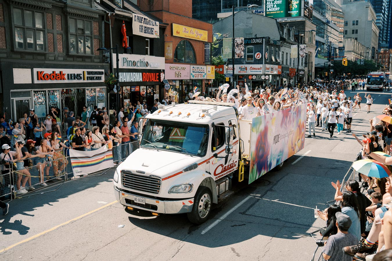 A vibrant pride parade float moves through a city street, surrounded by cheering spectators holding rainbow flags and umbrellas.