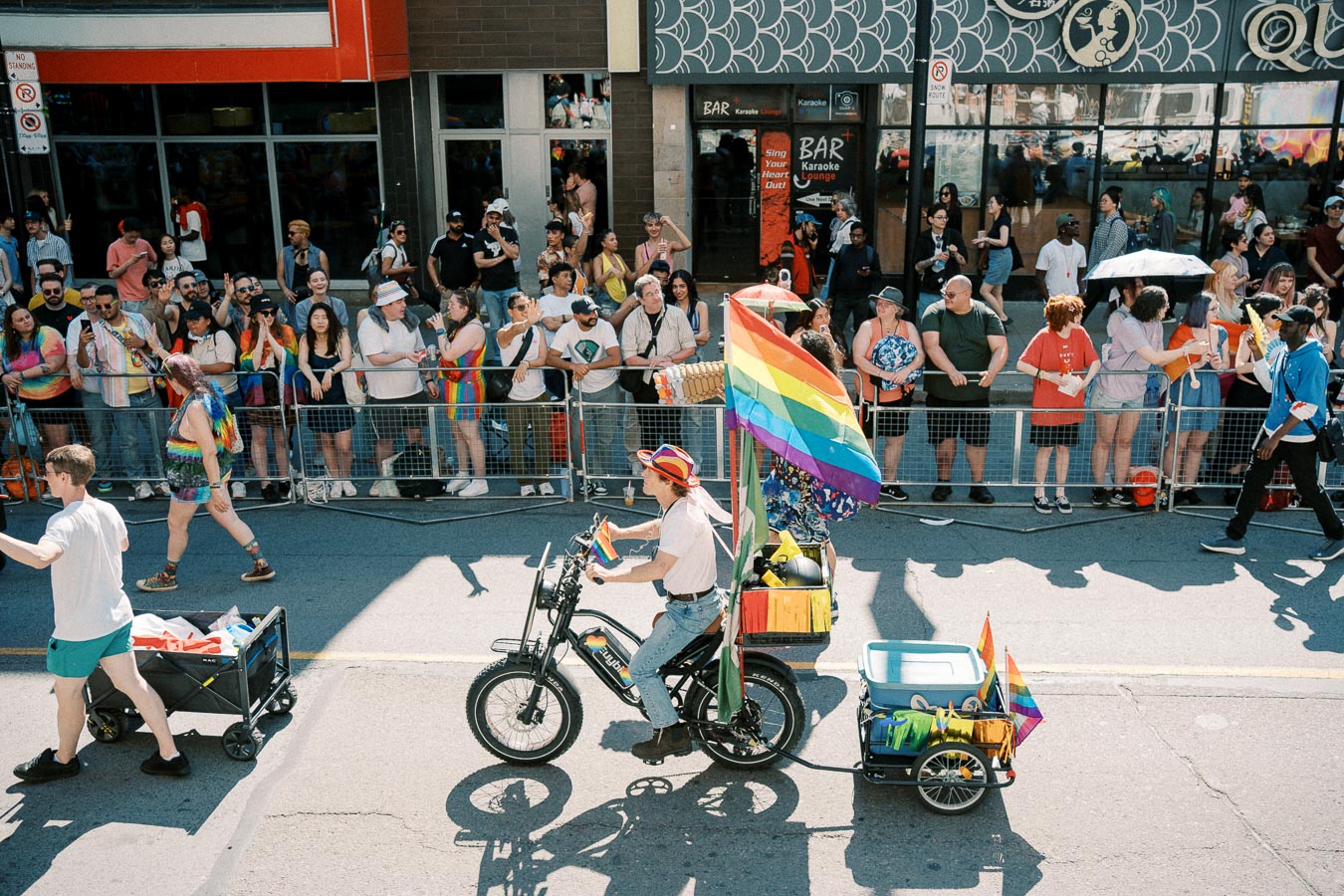 A vibrant scene from a pride parade featuring a person on a bicycle adorned with a rainbow flag, passing by a cheering crowd.