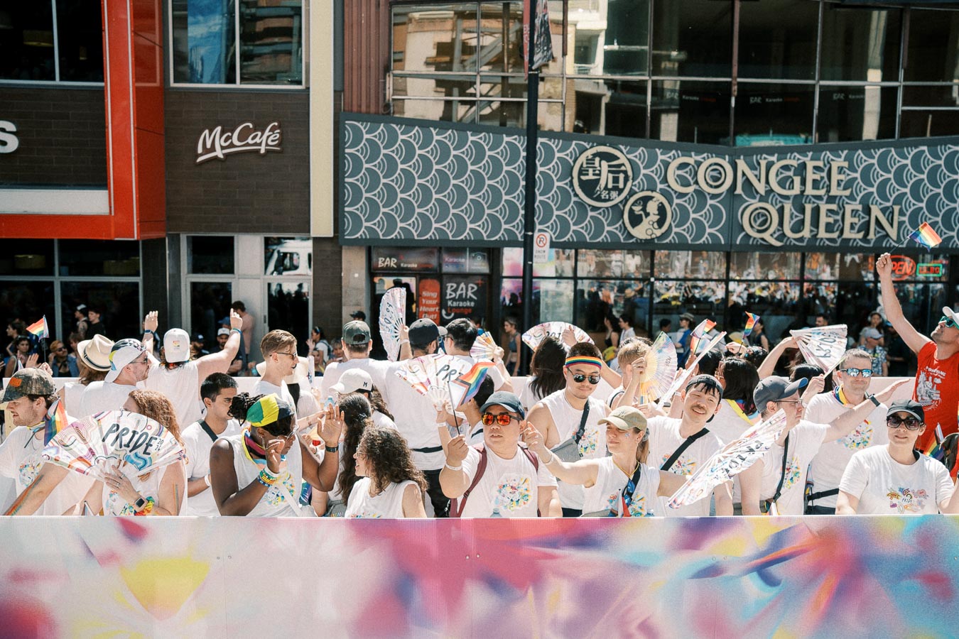 Participants celebrate at a pride parade, waving rainbow flags and fans, in front of a McCafé and Congee Queen restaurant.