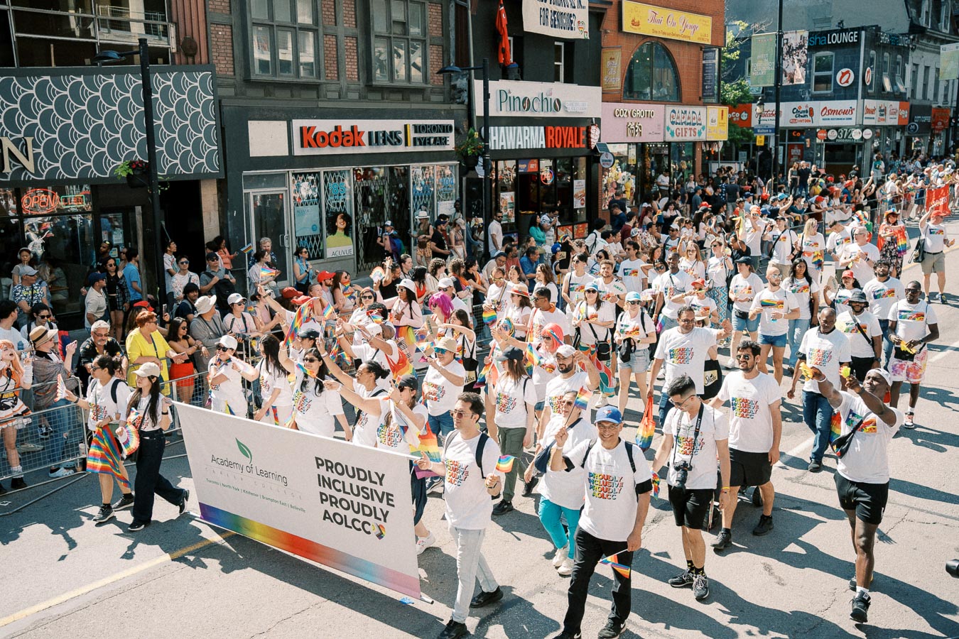 Participants in a pride parade march down a city street, celebrating diversity and inclusion with rainbow flags and matching T-shirts.