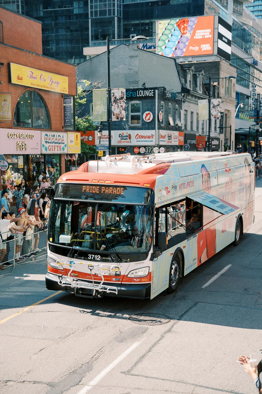 A bus decorated for the Pride Parade drives through a city street, surrounded by spectators. The bus displays 'Pride Parade' on its front, and the street is lined with shops and restaurants.
