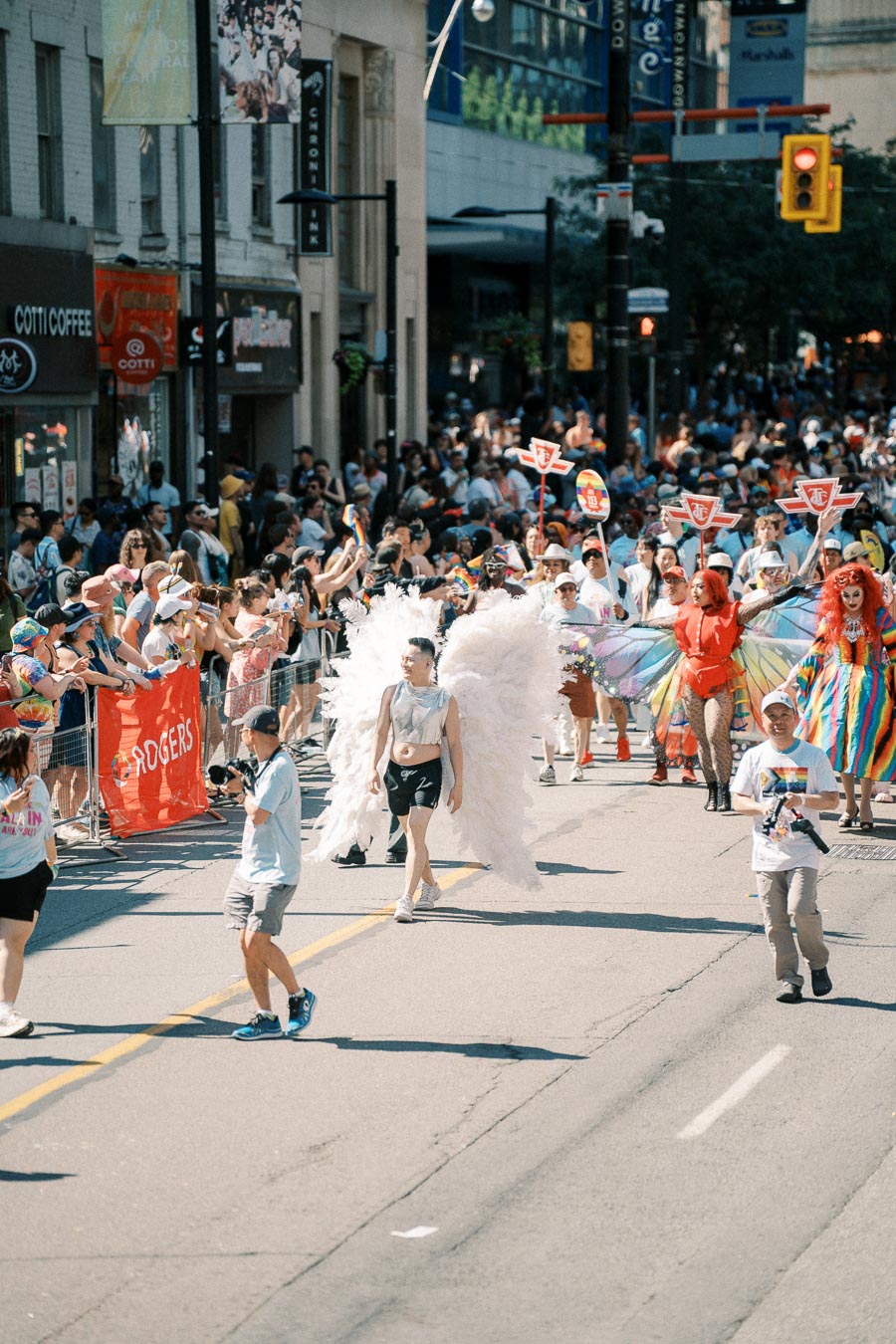 Participants in colorful costumes, including large white wings, march in a lively city parade with cheering spectators.