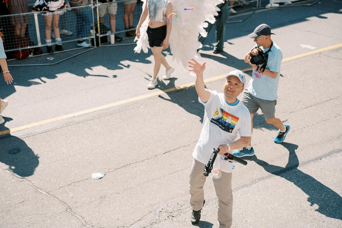A man waves during a parade, wearing a t-shirt with a rainbow design, while holding a camera. A person with angel wings and a photographer are seen in the background.