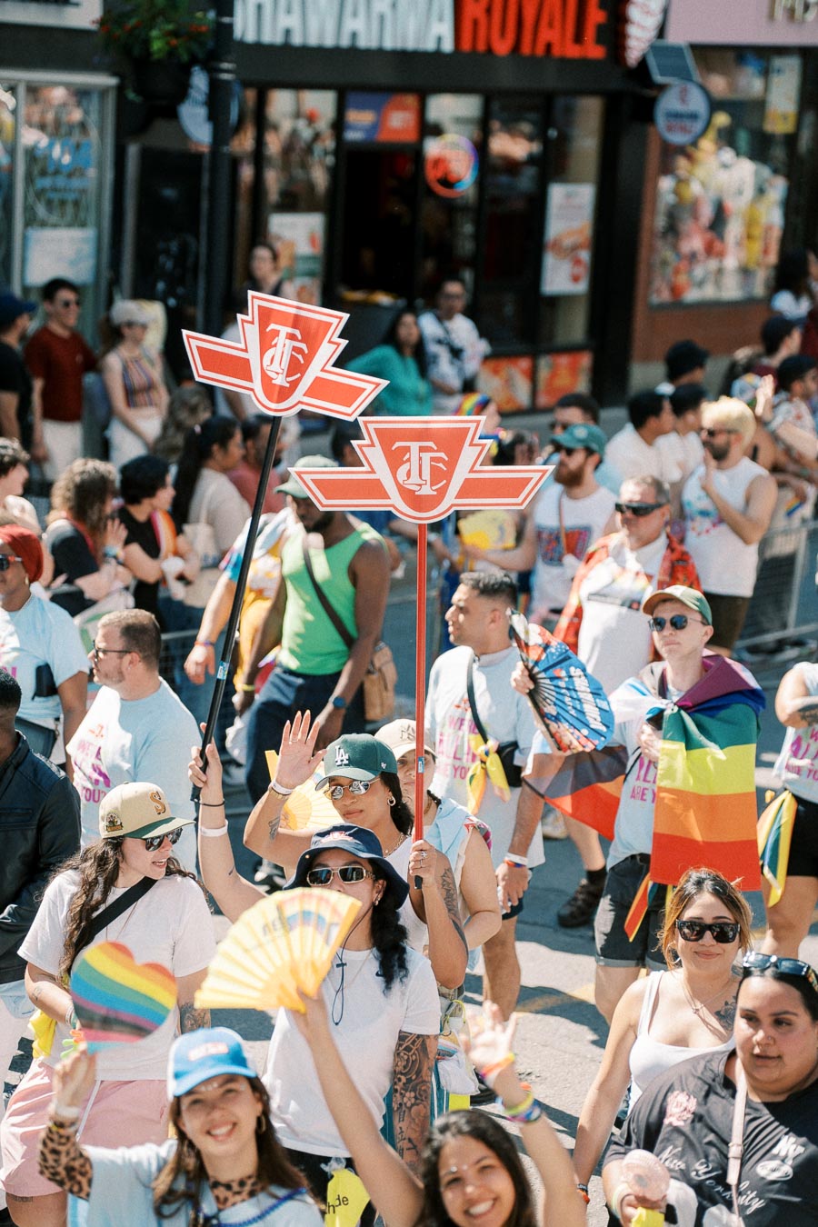 Participants in a parade hold signs with a red logo and wave rainbow fans, celebrating diversity and inclusion.