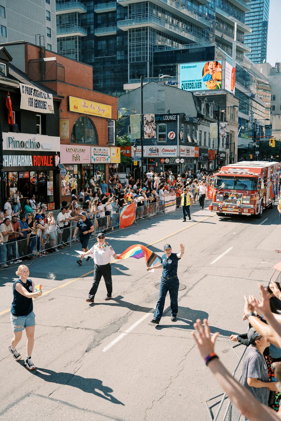 Participants in a pride parade wave rainbow flags as they walk down a city street, with a fire truck following behind. Spectators line the street, cheering and taking photos.