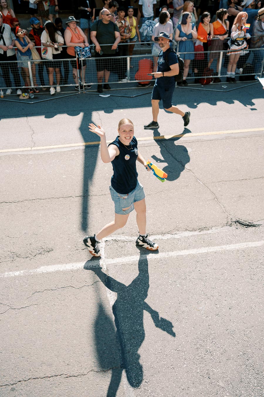 A person waves enthusiastically while holding a colorful water gun during a parade. They are walking on a street, wearing casual attire, and are surrounded by a crowd of onlookers behind a barrier.