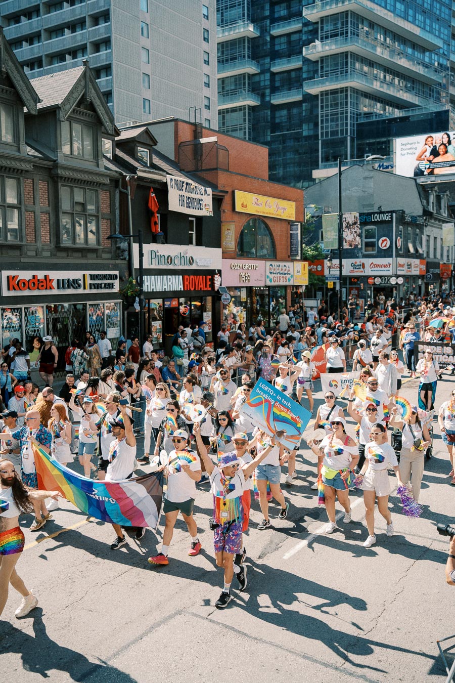 Participants in a pride parade march down a city street, celebrating with rainbow flags and banners, surrounded by onlookers and urban buildings.
