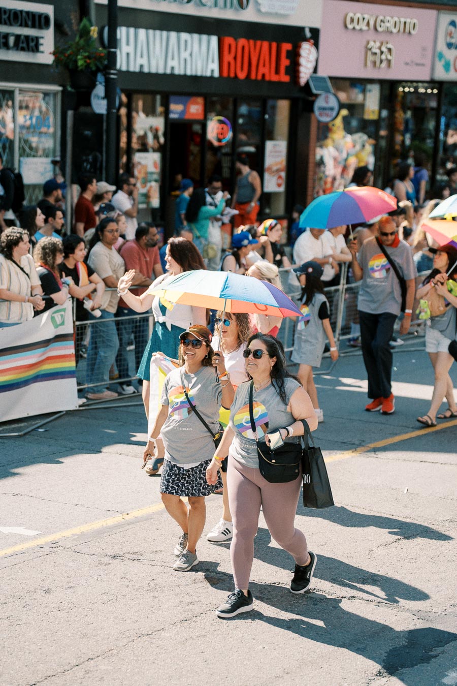 Participants in a pride parade walk down the street, holding colorful rainbow umbrellas and wearing shirts with rainbow designs. Spectators line the sidewalk, watching the procession.