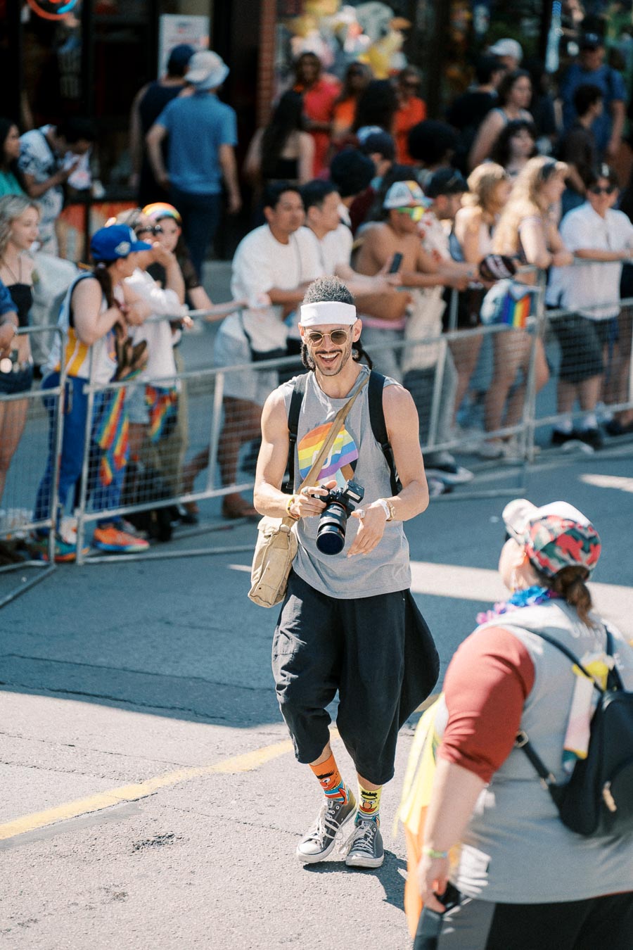 A photographer captures moments at a pride parade, wearing a rainbow-themed outfit and smiling amidst the lively crowd.
