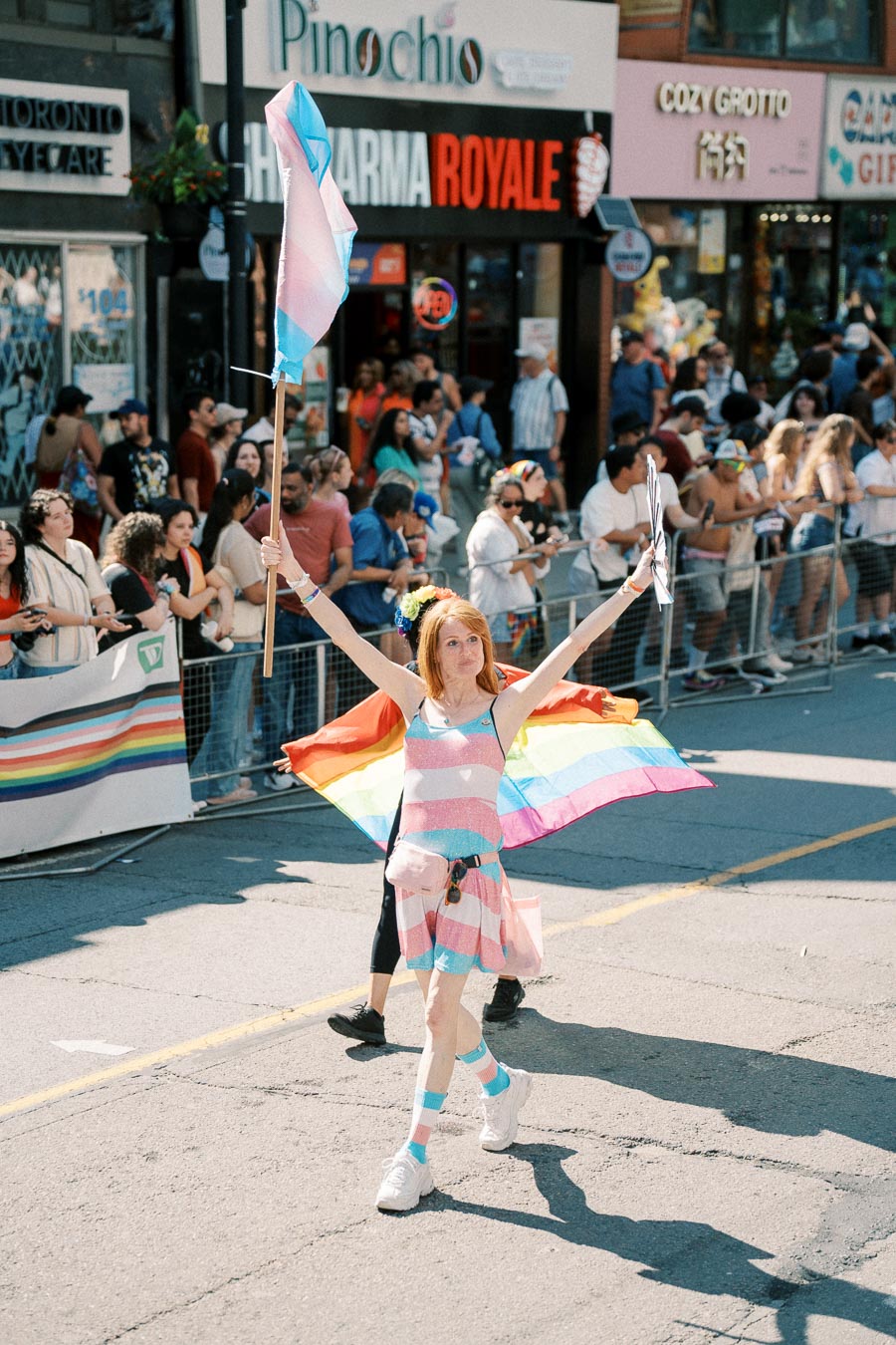 A participant in a pride parade holds a transgender pride flag and wears a rainbow cape, celebrating diversity and inclusion.