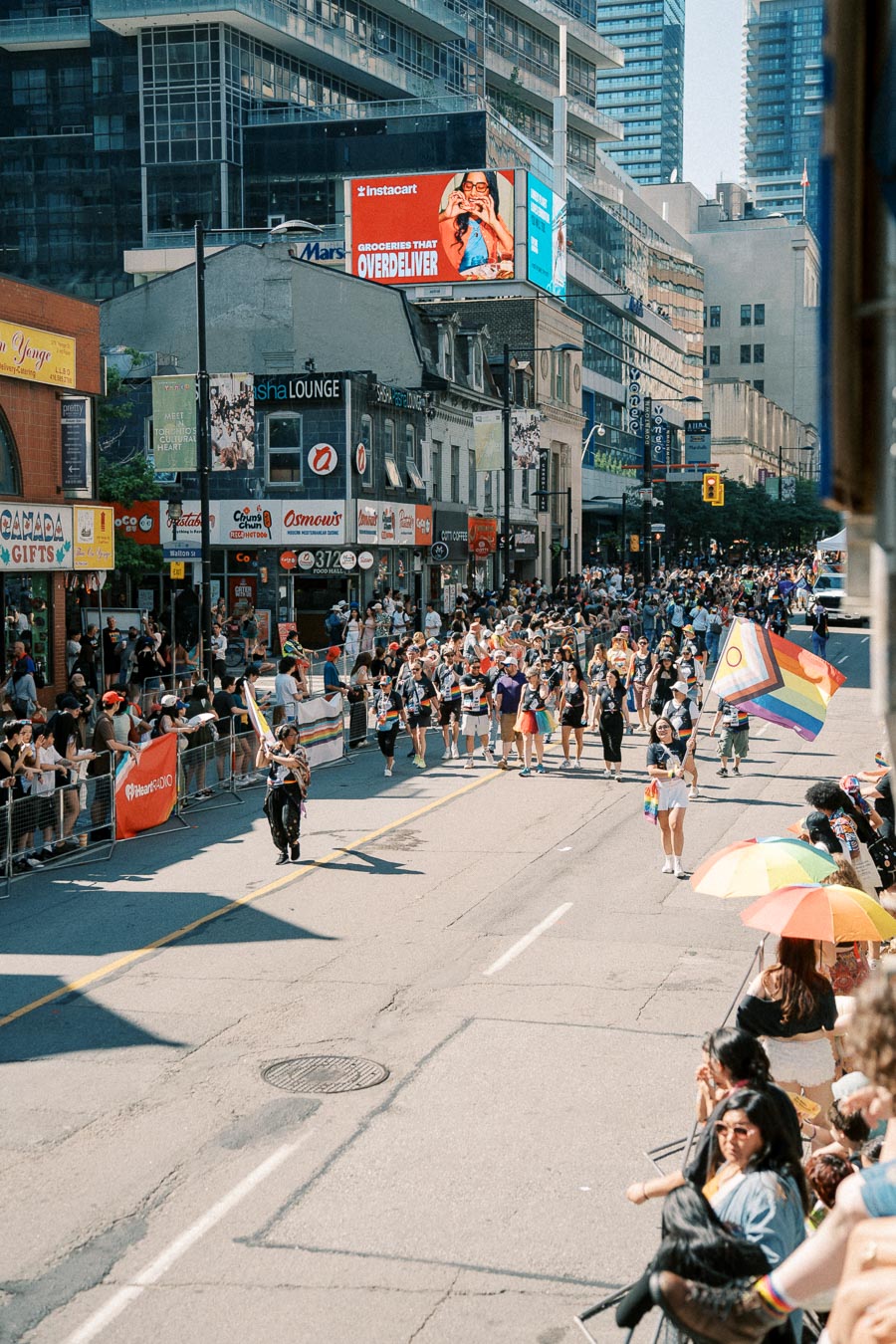 A lively parade takes place on a city street, featuring participants with rainbow flags and a crowd of spectators. The scene is set against a backdrop of urban buildings and colorful advertisements.