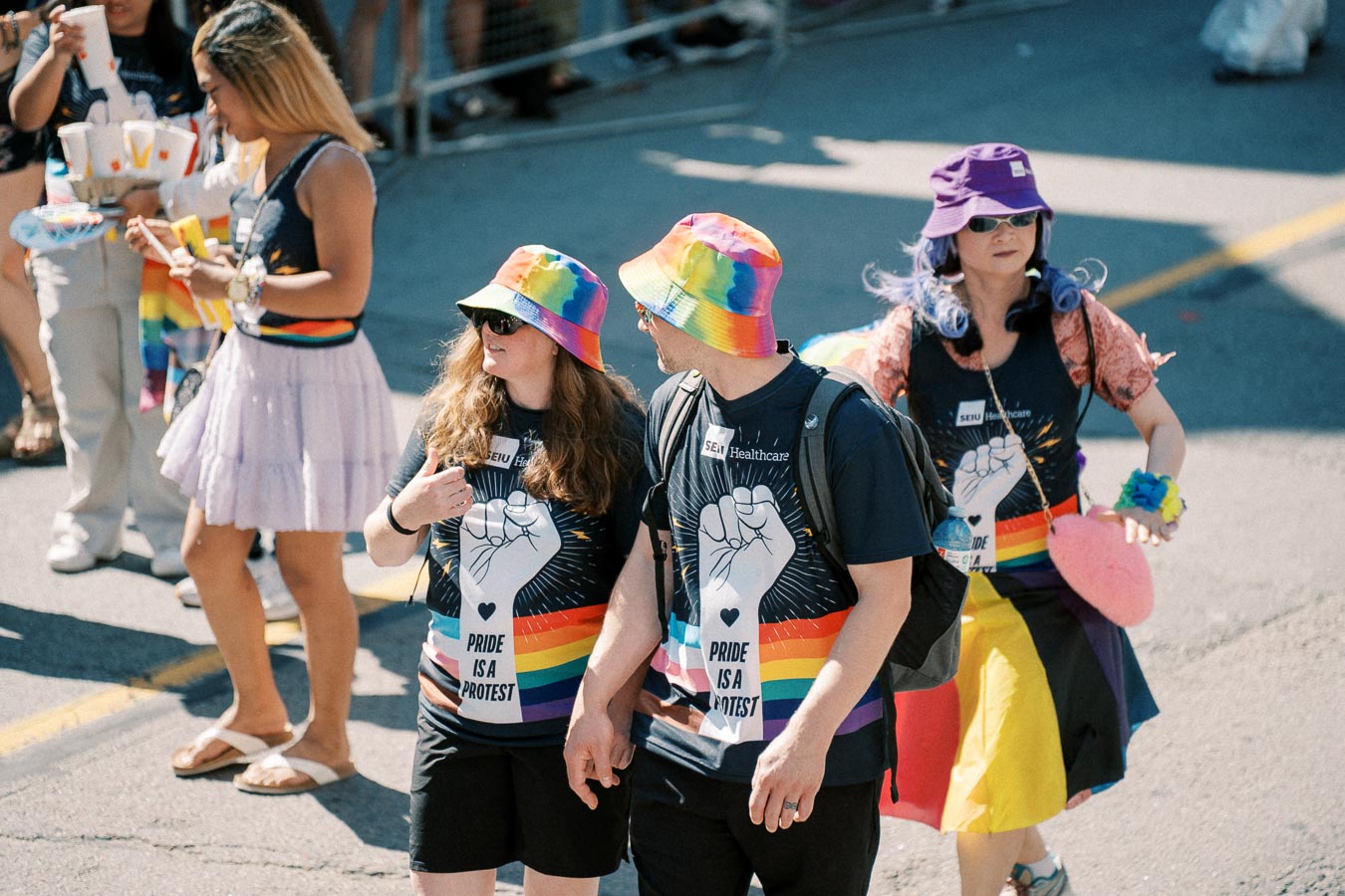 Participants in a pride parade wearing rainbow-themed hats and shirts with the message 'Pride is a Protest.'