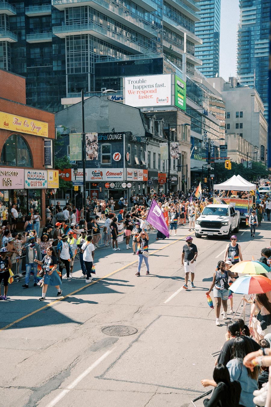 A lively street parade featuring participants with rainbow flags and banners, surrounded by spectators and urban buildings.