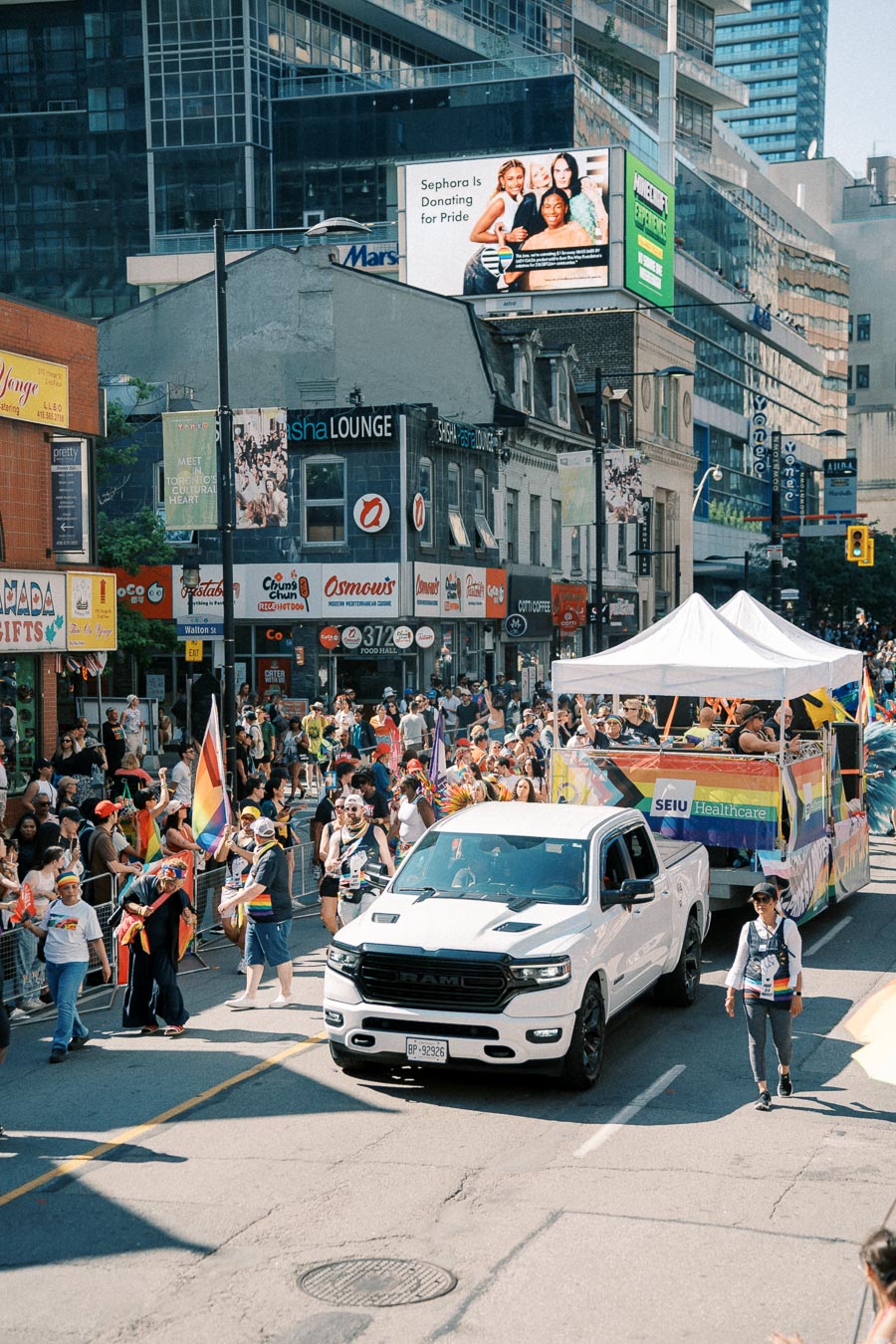 A vibrant pride parade takes place in a city, featuring a white truck towing a colorful float. Participants walk alongside, waving rainbow flags, while spectators line the street. Urban buildings and advertisements form the backdrop.