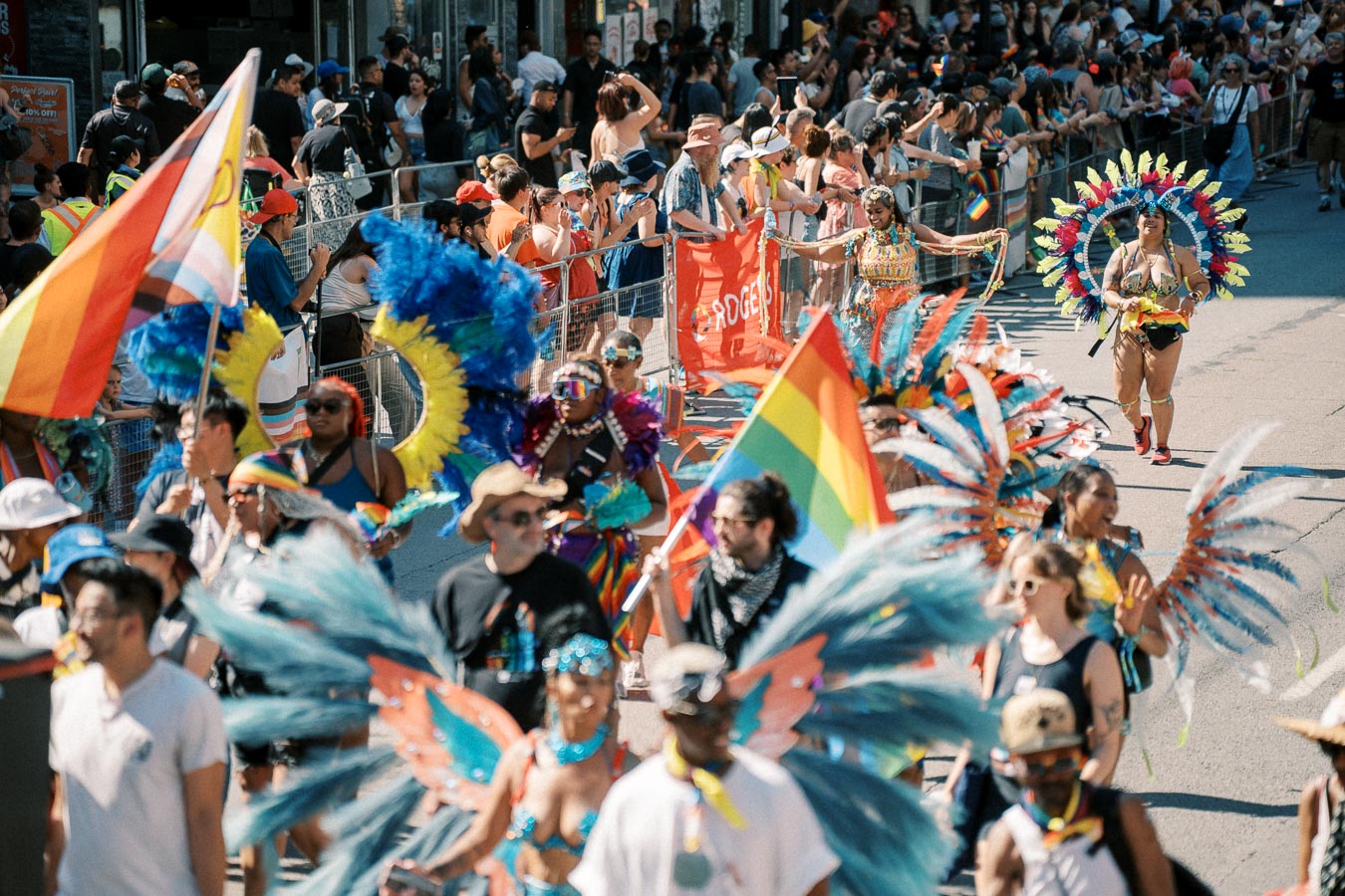Participants in colorful costumes and rainbow flags march in a lively parade, watched by a crowd of spectators.