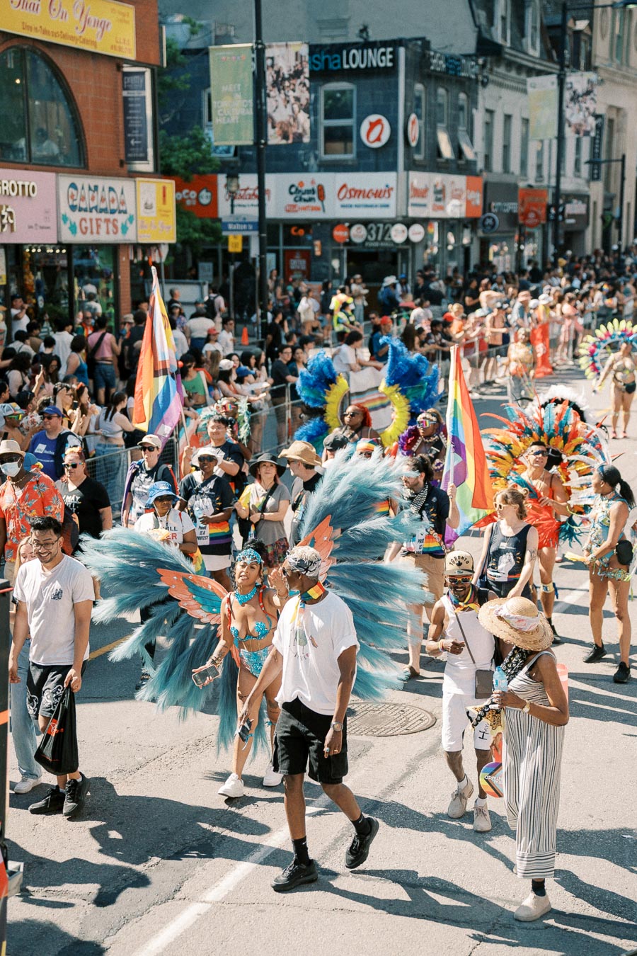 Participants in colorful costumes and rainbow flags march in a lively parade, with spectators lining the city street.