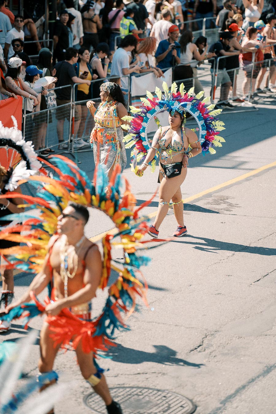Participants in vibrant, feathered costumes march in a lively street parade, with onlookers gathered behind barriers.