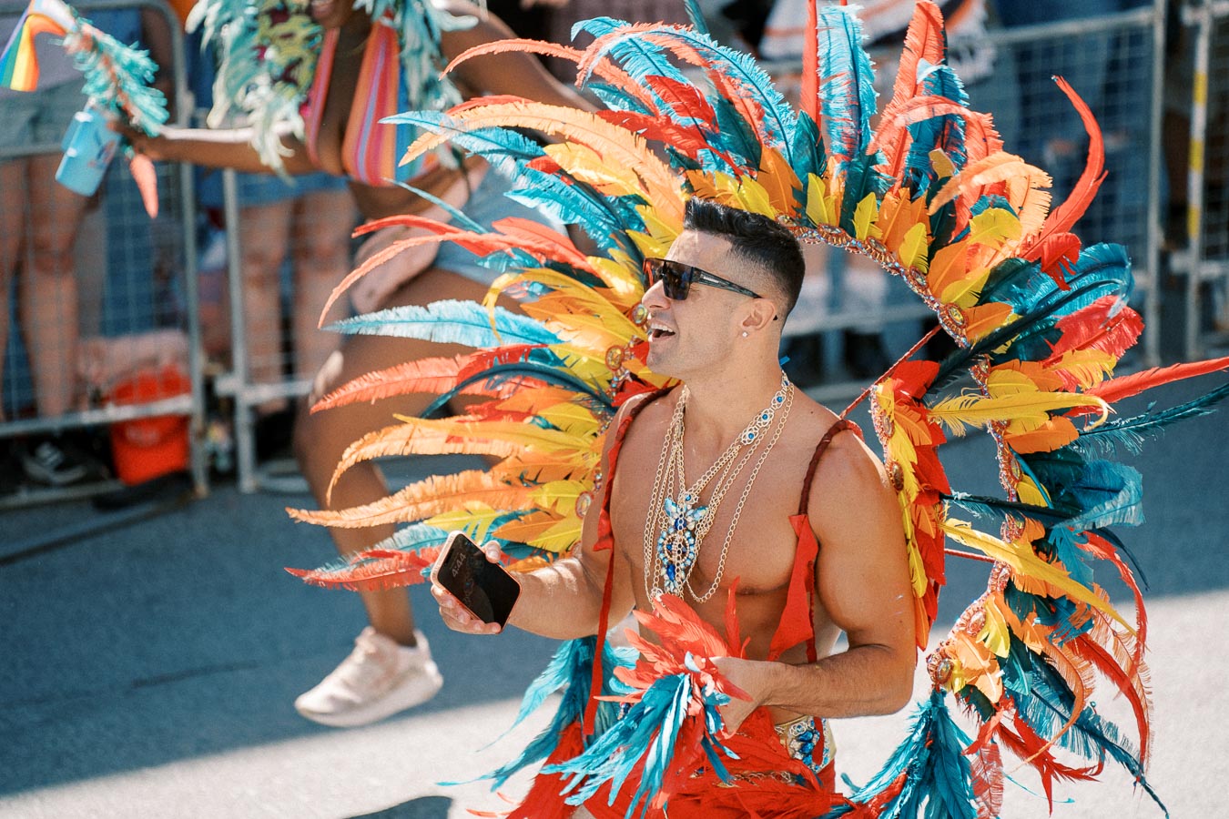 A participant in a vibrant feathered costume enjoys a parade, holding a phone and smiling.