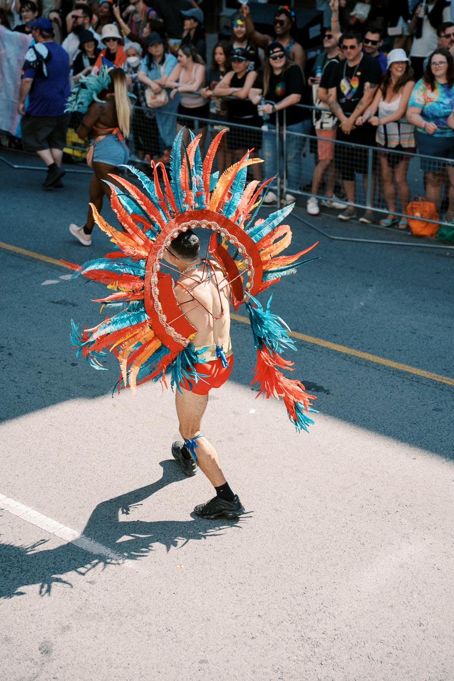 A vibrant parade scene featuring a person in a colorful feathered costume, walking along a street lined with spectators. The costume is adorned with red, orange, and blue feathers, creating a striking visual display.