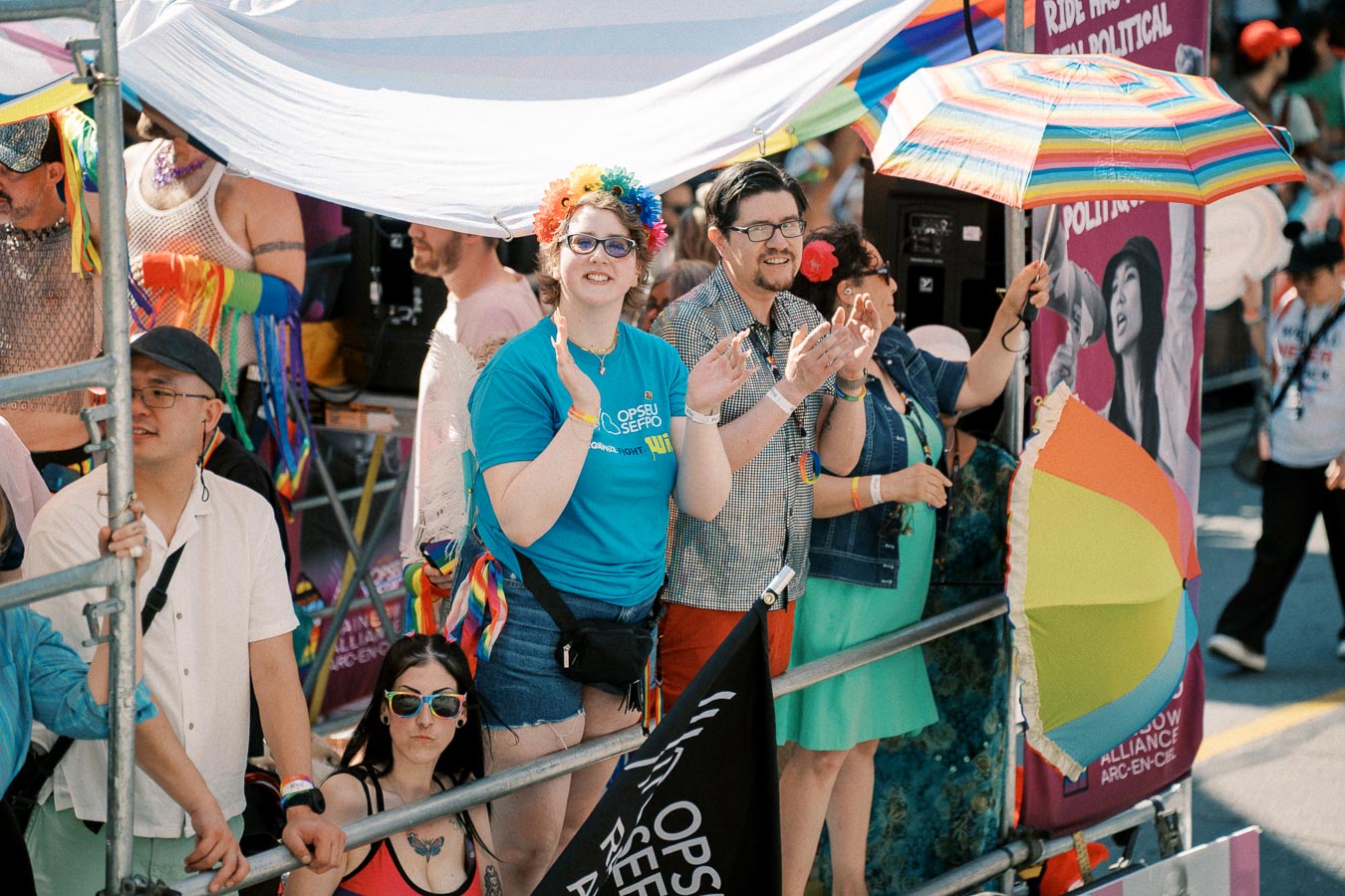 Participants at a pride parade celebrate with colorful attire and rainbow-themed accessories, including a rainbow umbrella and flower crown.