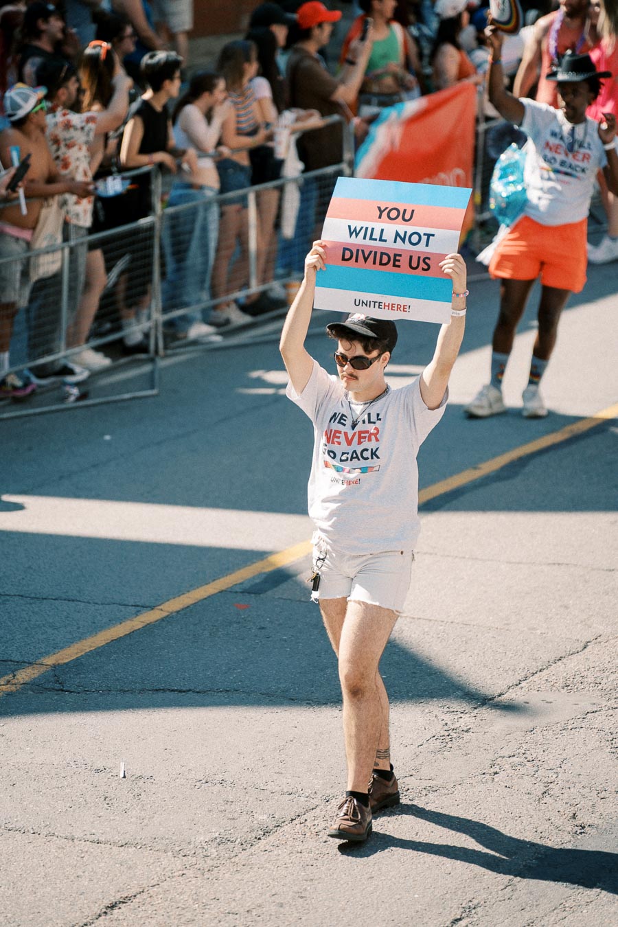 A person participates in a parade, holding a sign that reads 'You Will Not Divide Us' while wearing a t-shirt with a similar message. The event is attended by a diverse crowd.