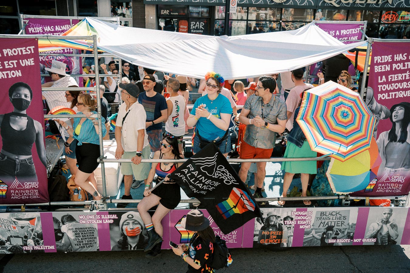 A vibrant parade float features people celebrating with rainbow-themed decorations and protest signs advocating for LGBTQ+ rights and social justice.