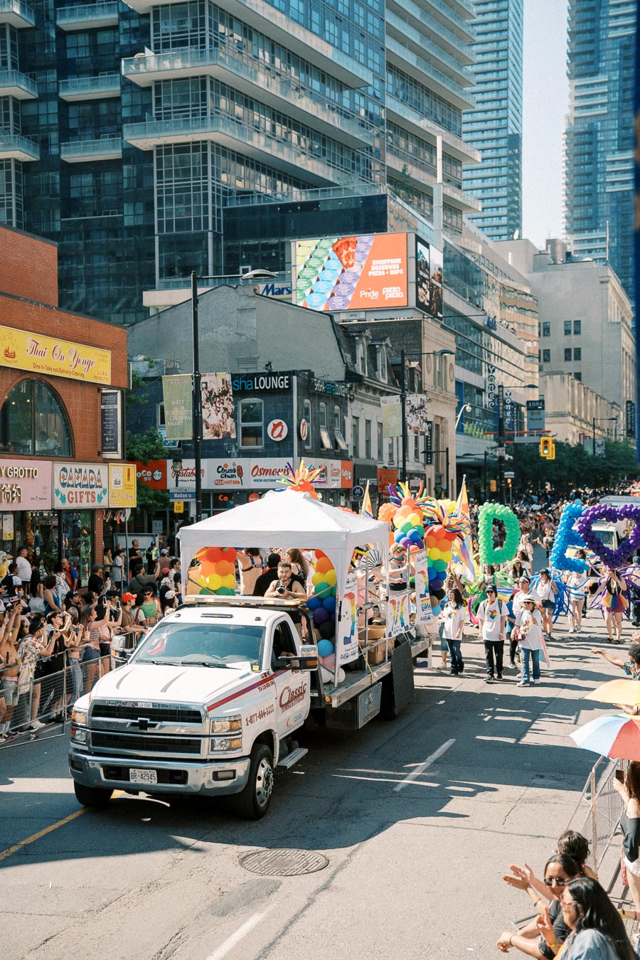 A parade float decorated with rainbow colors moves down a city street during a pride event, surrounded by cheering spectators and tall buildings.