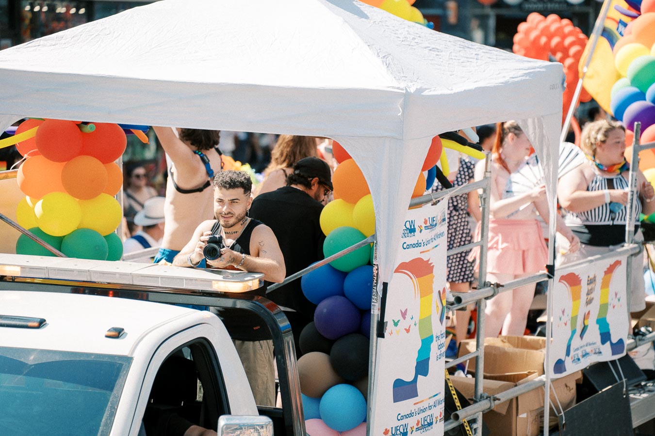 A vibrant parade float adorned with rainbow balloons and banners, featuring a person with a camera capturing the lively atmosphere.