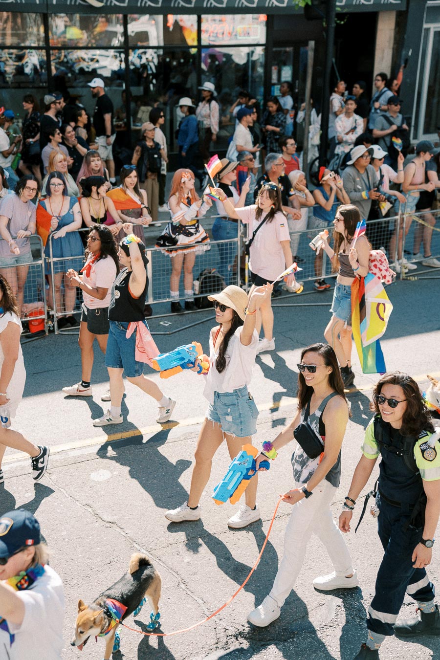Participants in a vibrant parade march down the street, waving rainbow flags and carrying water guns, while a cheerful crowd looks on. A dog joins the festivities, adorned with a colorful bandana.