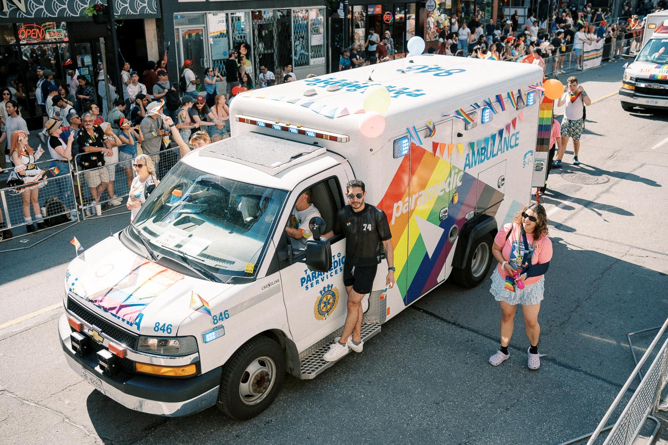 A rainbow-themed ambulance participates in a parade, adorned with colorful decorations and balloons, as people gather to watch the event.