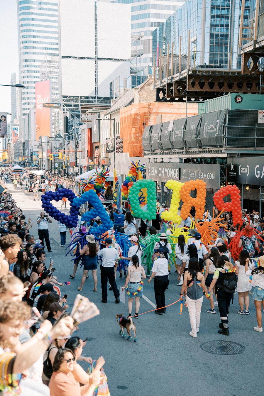A lively pride parade with colorful balloon letters spelling 'PRIDE' and a cheering crowd in an urban setting.