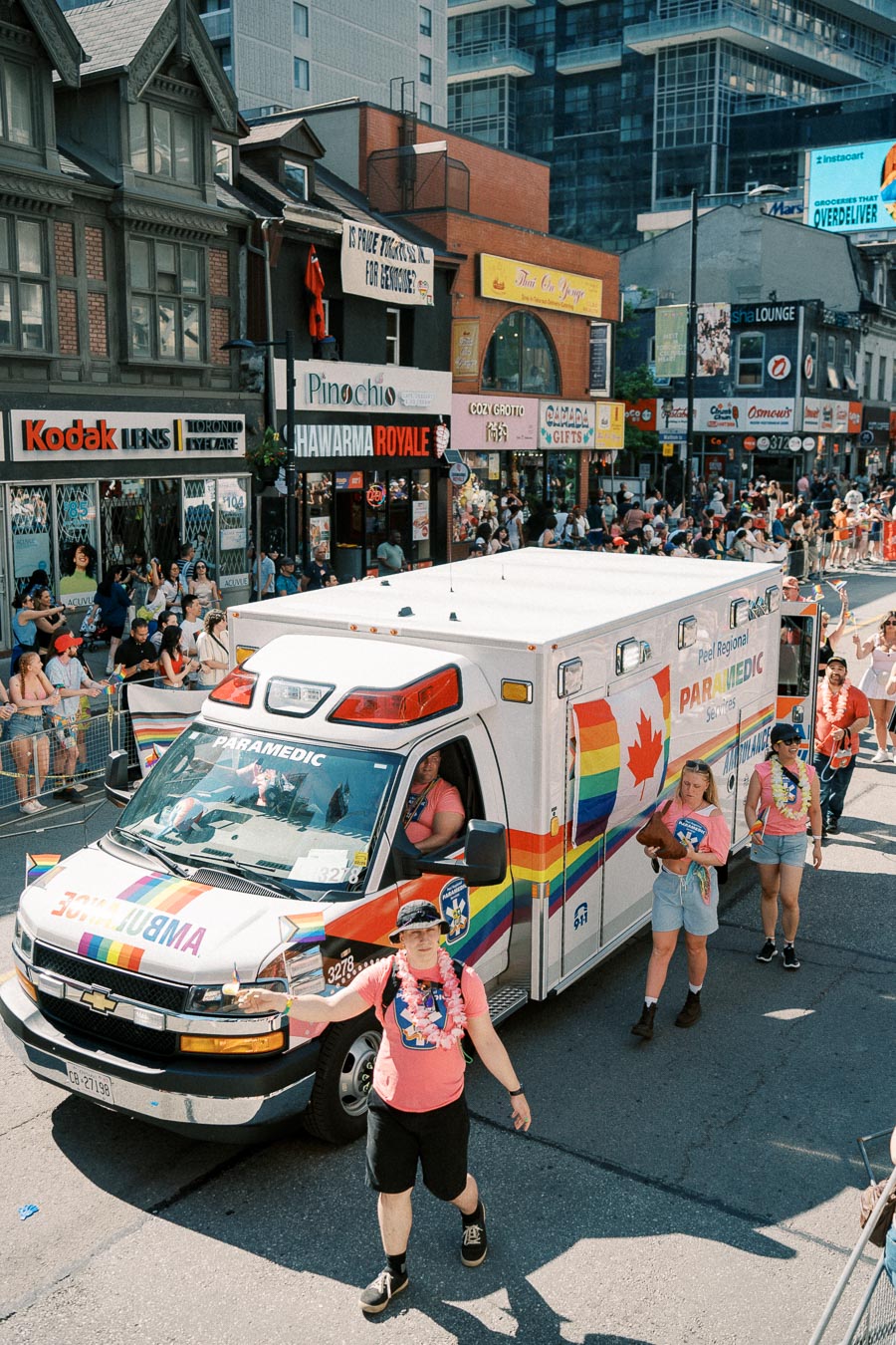 A rainbow-themed ambulance participates in a parade, with people in pink shirts walking alongside, celebrating diversity and inclusion.