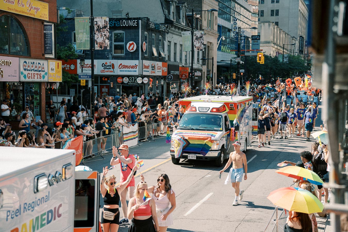 A colorful parade featuring a rainbow-decorated ambulance and participants celebrating with flags, observed by a large crowd on a city street.