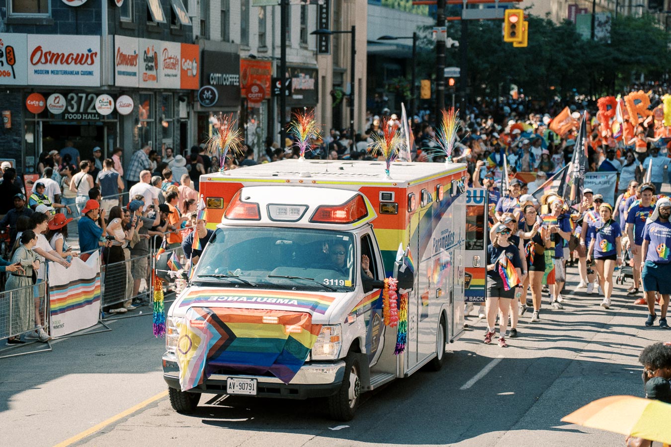 A rainbow-decorated ambulance participates in a pride parade, surrounded by a lively crowd and marchers.