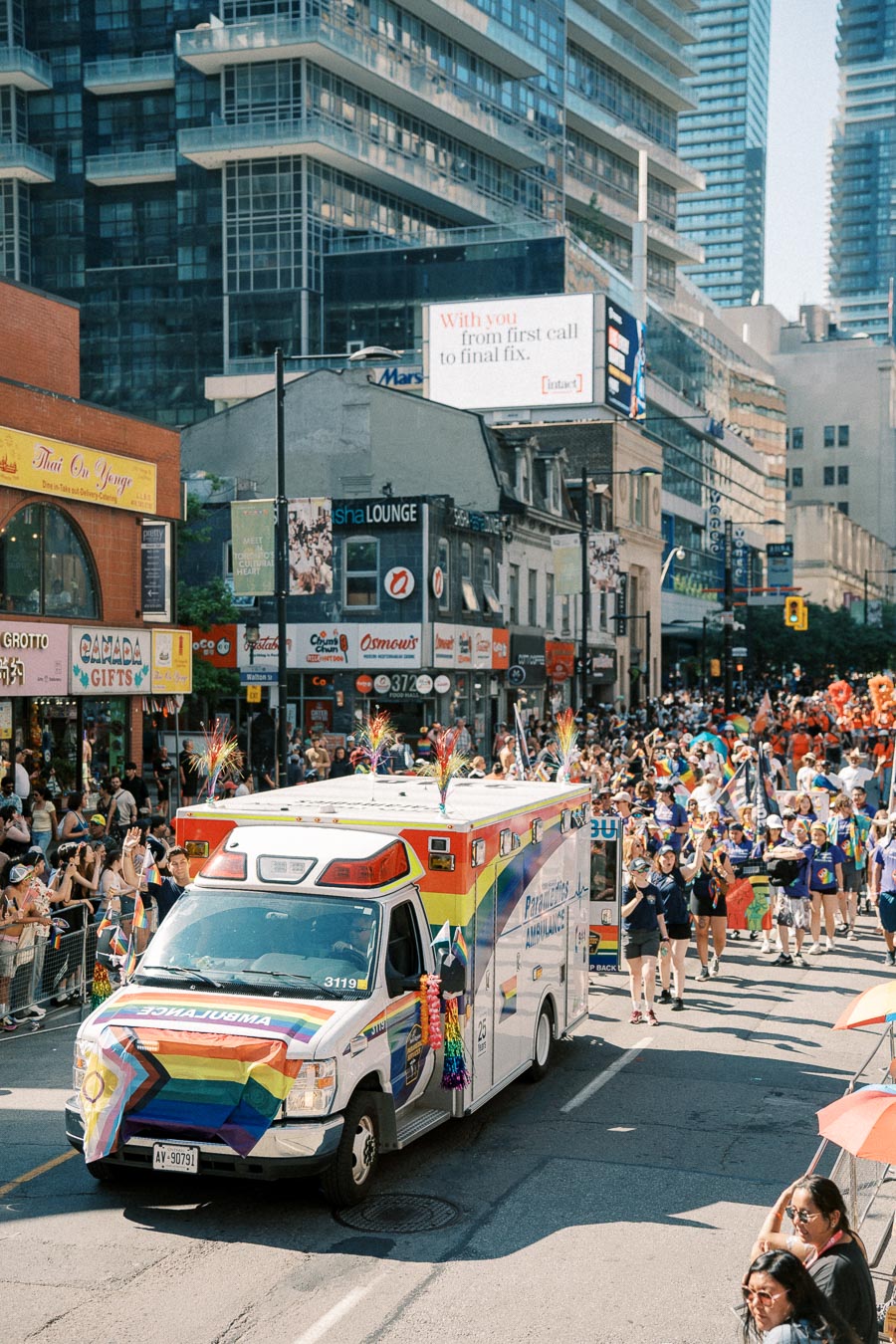 A vibrant parade featuring a rainbow-decorated ambulance and participants marching down a busy city street, surrounded by spectators and tall buildings.