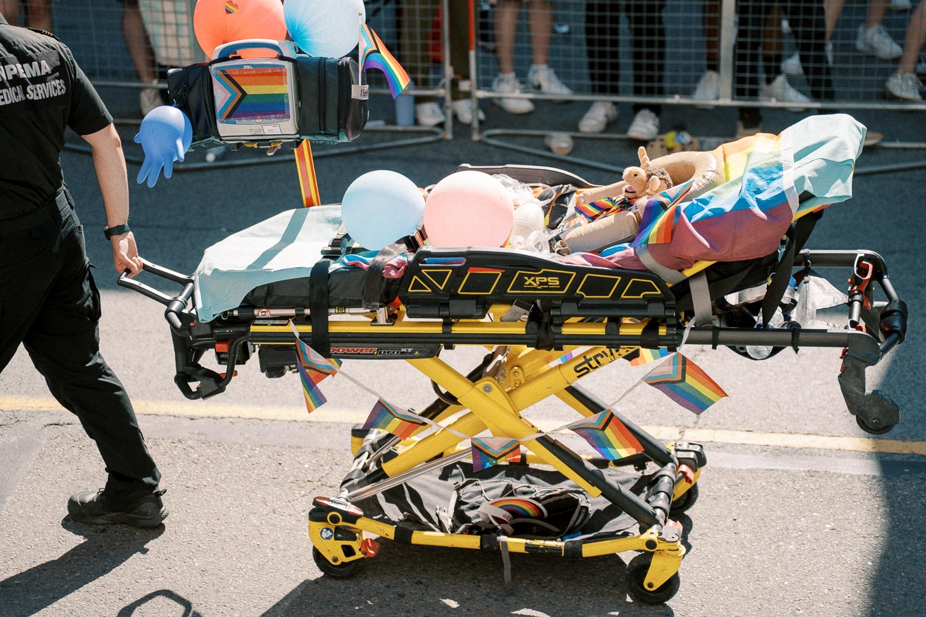 A stretcher adorned with rainbow flags, balloons, and a stuffed animal is being pushed by a person in a parade, symbolizing support for LGBTQ+ pride.