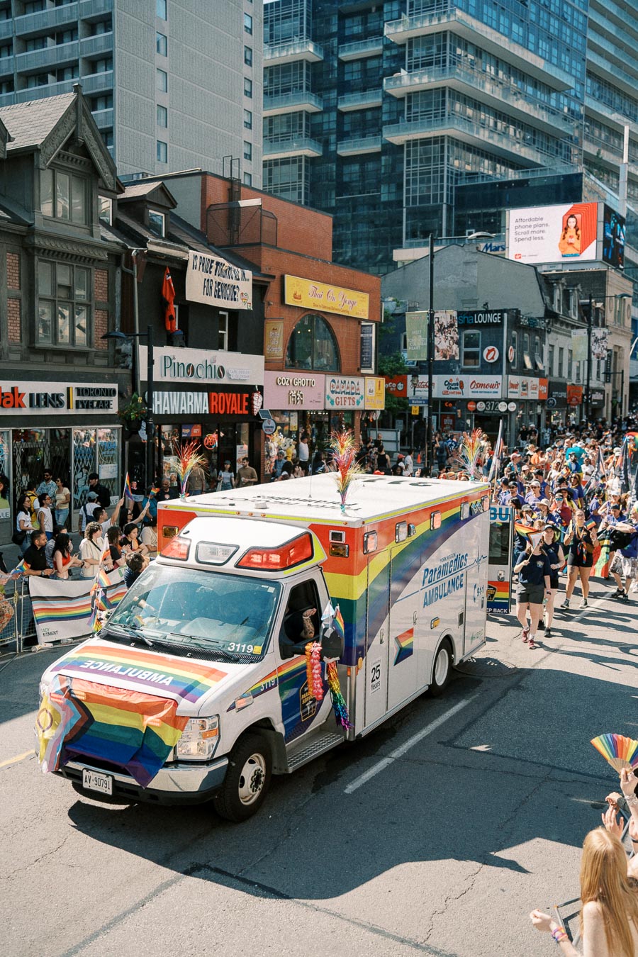 A rainbow-themed ambulance drives through a pride parade, surrounded by cheering crowds and city buildings.