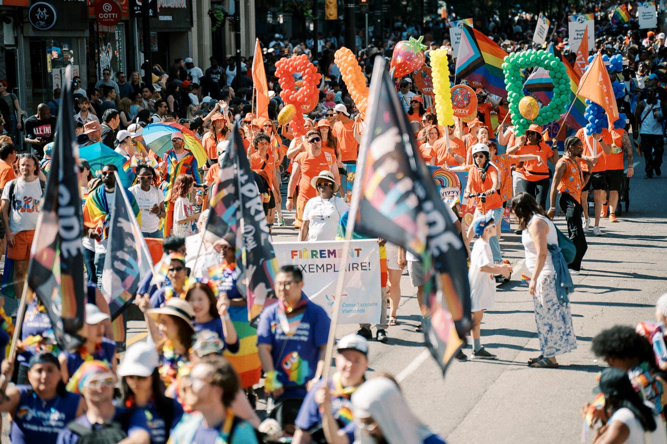 Participants in a colorful Pride parade march down the street, holding rainbow flags and balloons spelling 'PRIDE'.