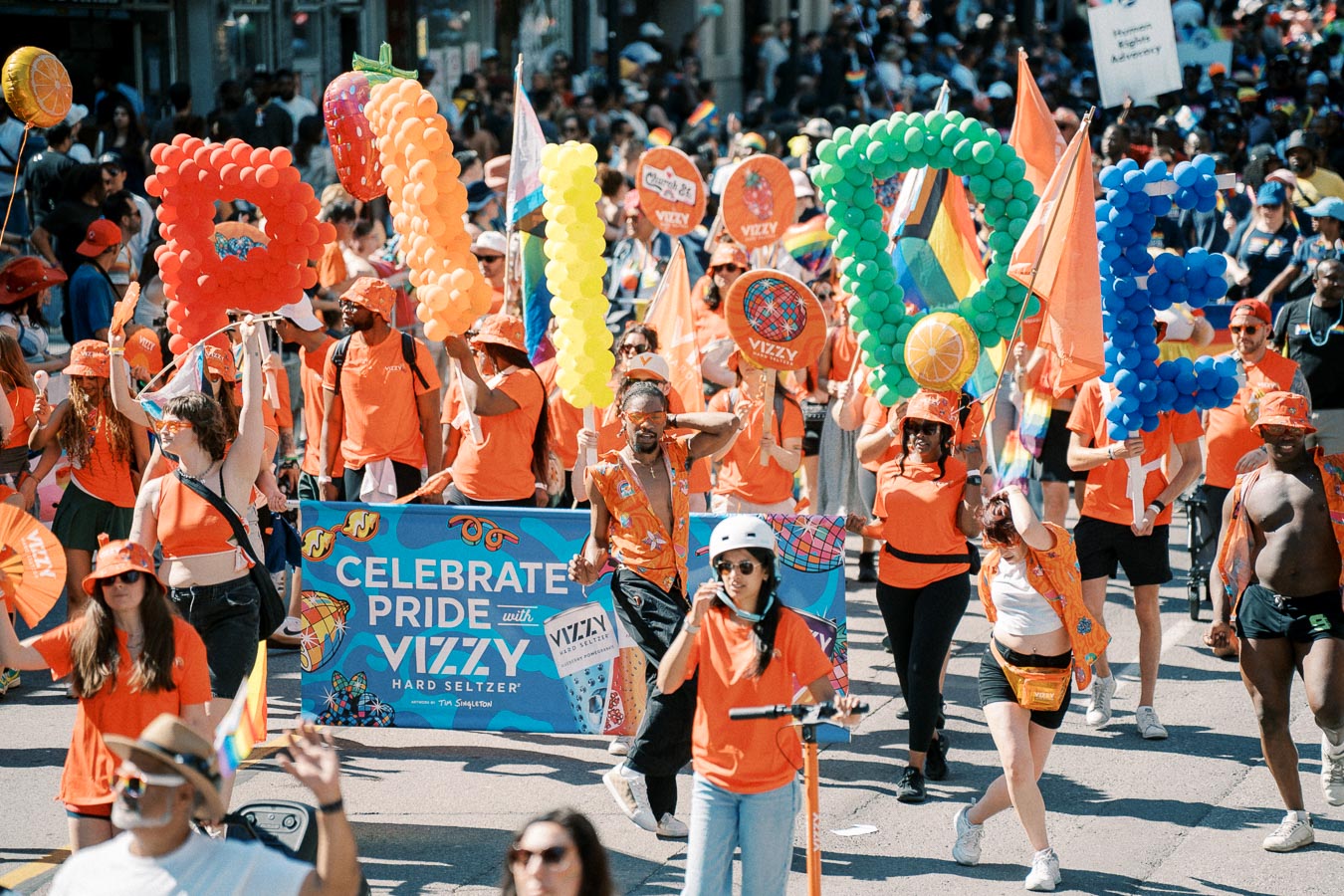 Participants at a Pride parade hold colorful balloon letters spelling 'PRIDE', celebrating with banners and vibrant outfits.