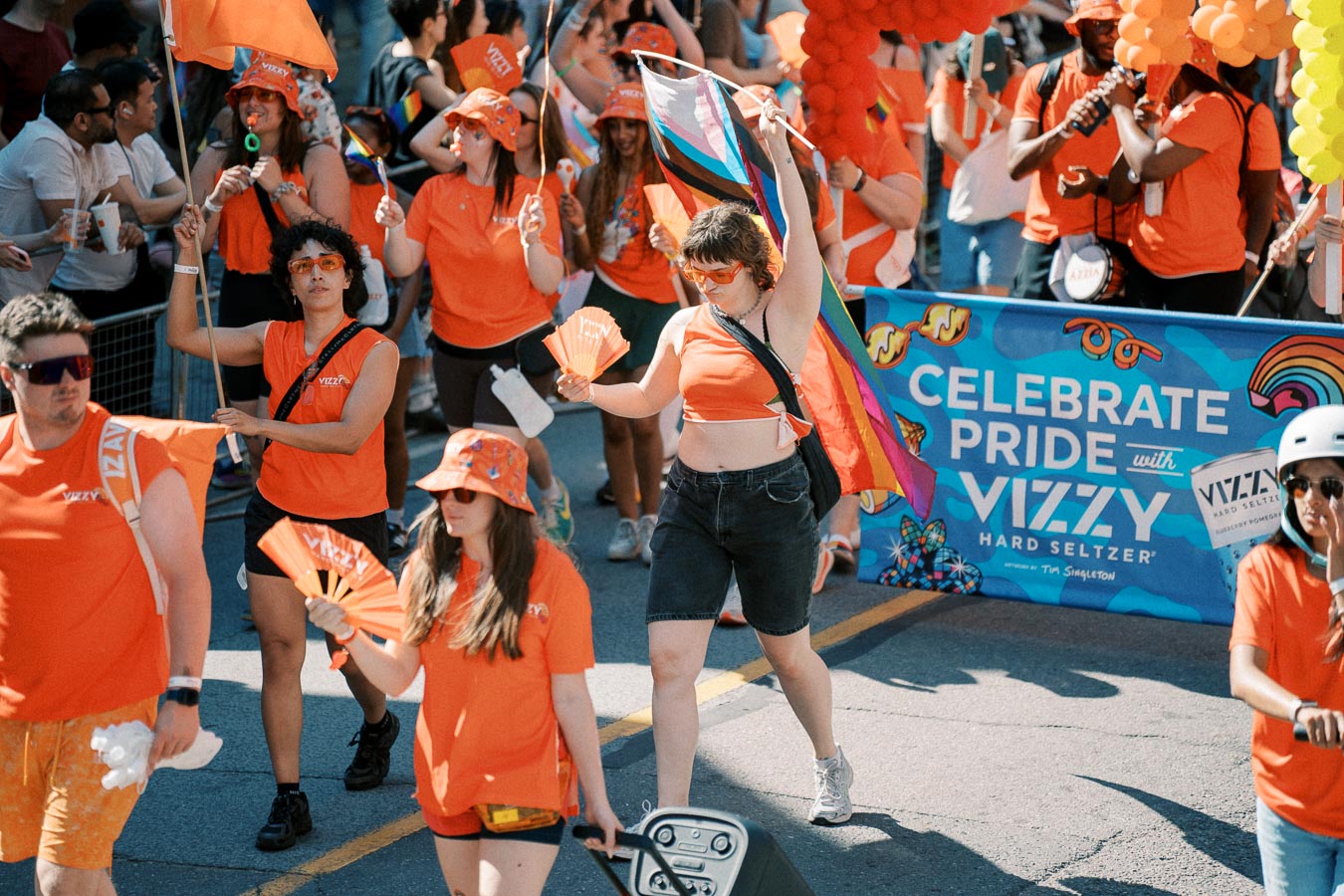 Participants in a Pride parade wearing orange outfits and holding flags and fans, celebrating with a banner promoting Vizzy Hard Seltzer.