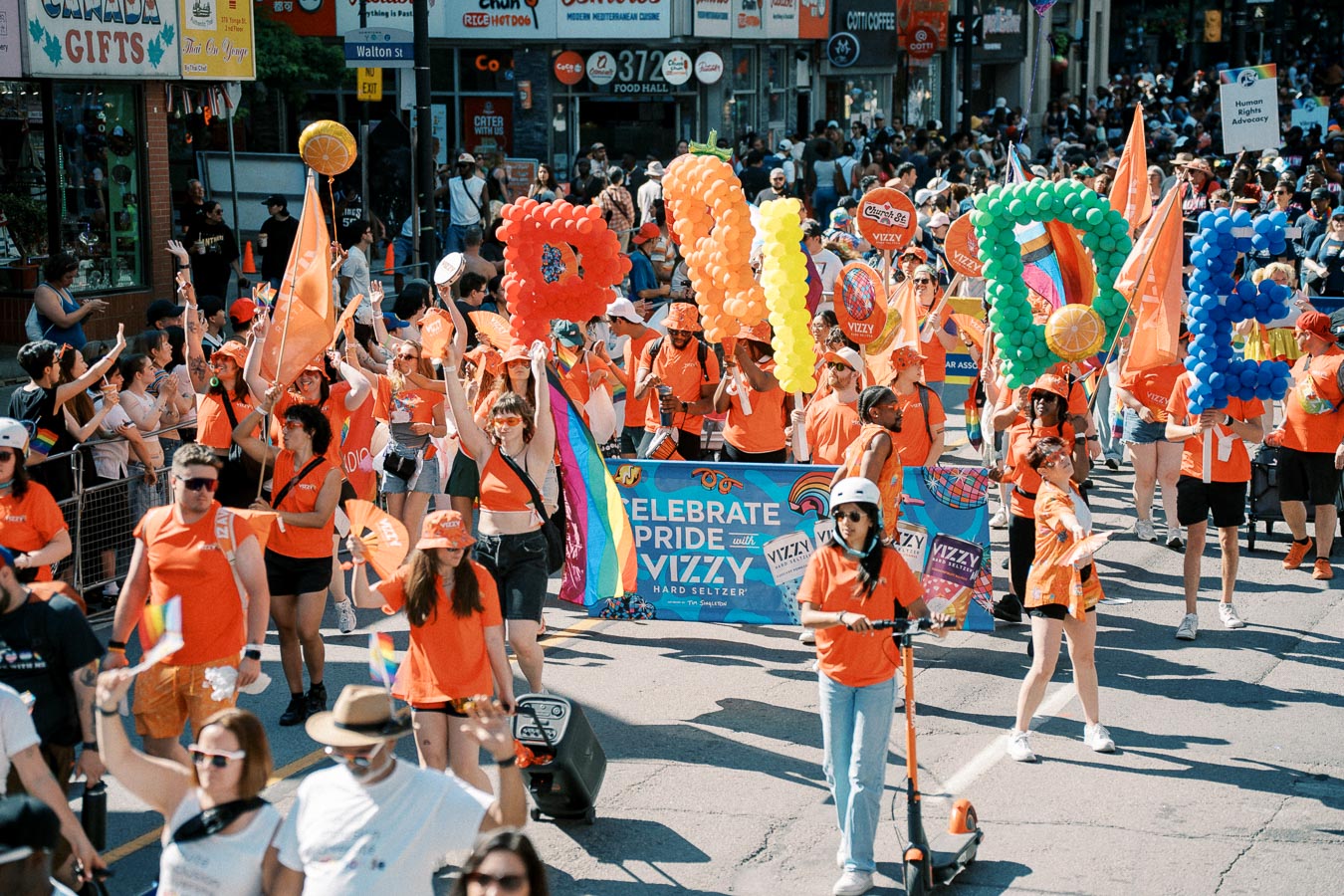 Participants in a Pride parade wearing orange shirts and holding colorful balloon letters spelling 'PRIDE'. The event is lively with rainbow flags and a banner promoting Pride with Vizzy Hard Seltzer.