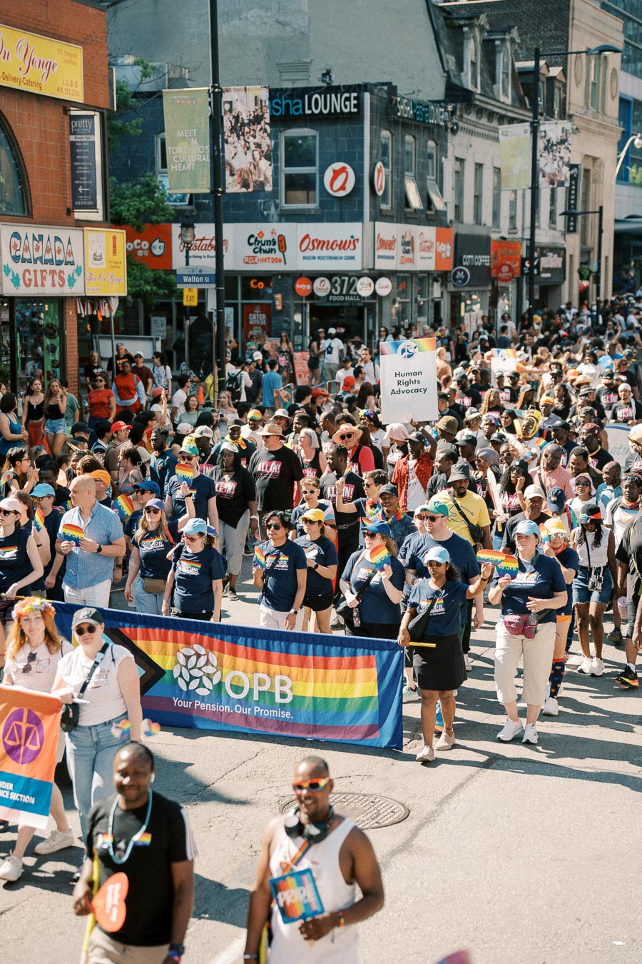 Participants march in a Pride parade, holding rainbow flags and banners, on a bustling city street lined with shops and restaurants.