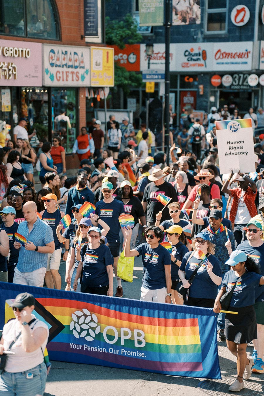 Participants in a pride parade march with rainbow flags and a banner reading 'OPB Your Pension. Our Promise.'