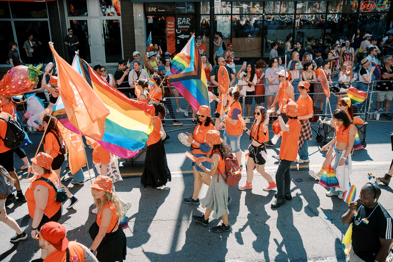 Participants in a pride parade march down the street, wearing orange attire and carrying rainbow flags, celebrating diversity and inclusion.