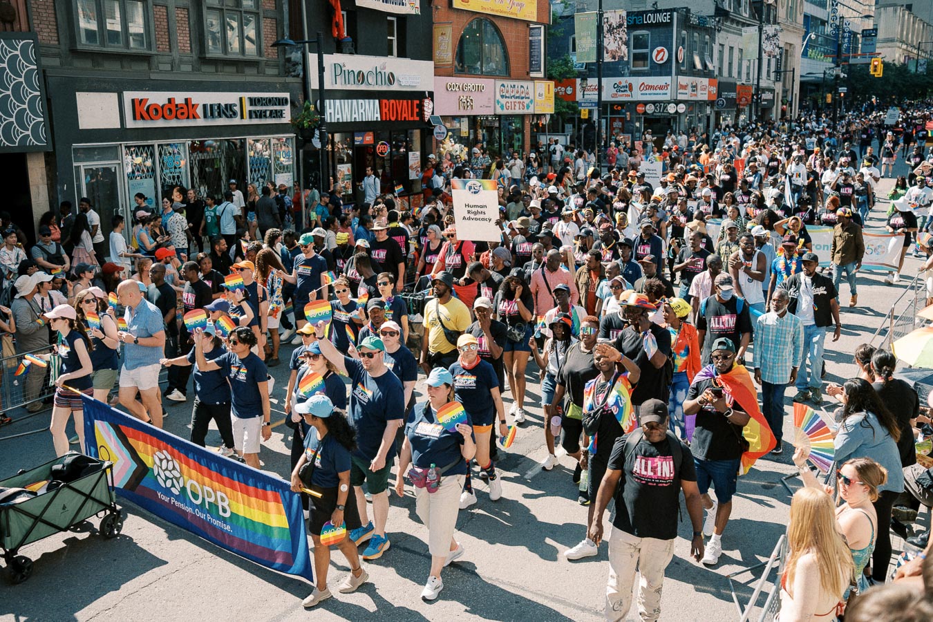 Participants in a pride parade march down a bustling city street, waving rainbow flags and carrying banners advocating for human rights.