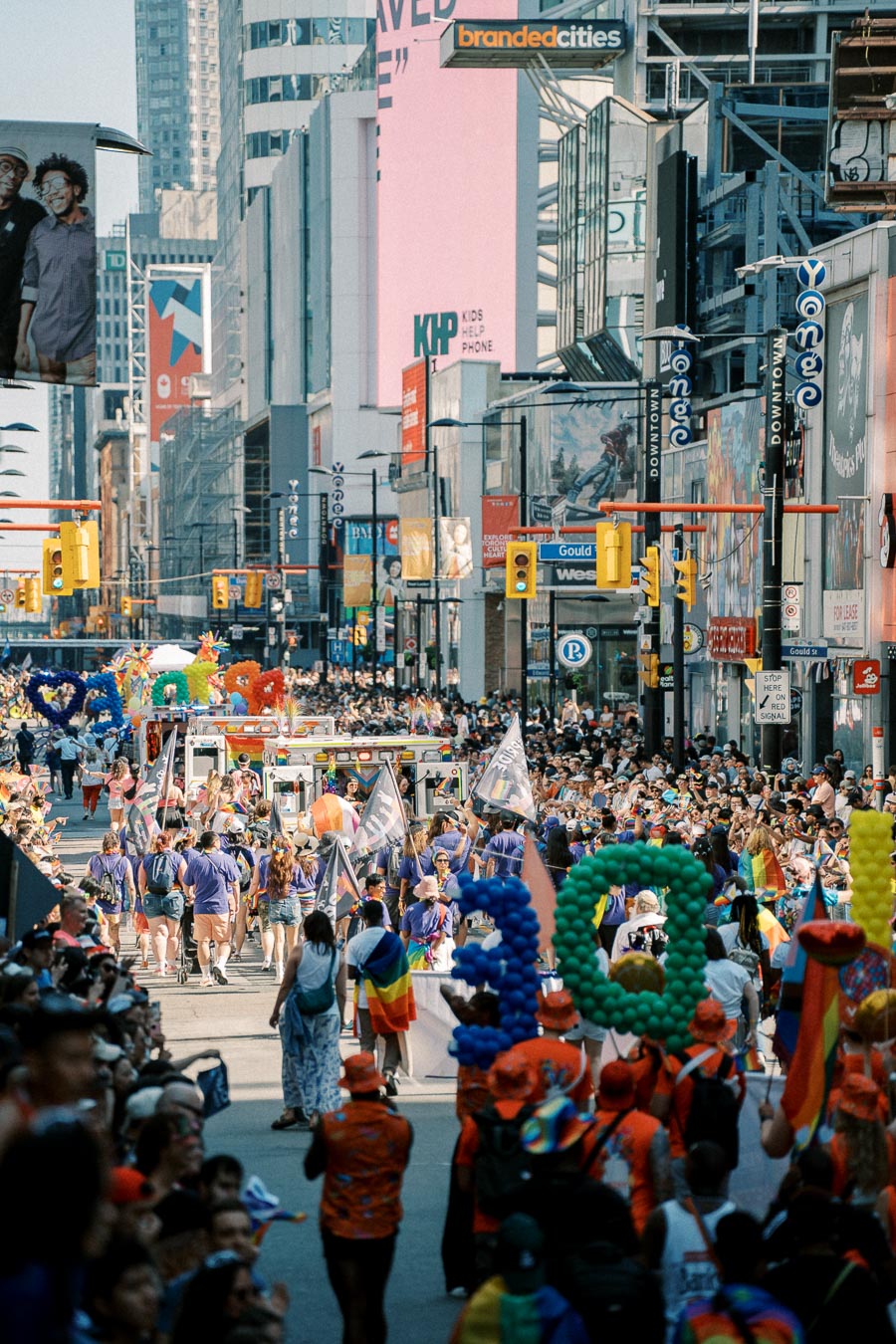 A lively parade fills a bustling city street, featuring participants in colorful attire and balloons, surrounded by tall buildings and vibrant advertisements.