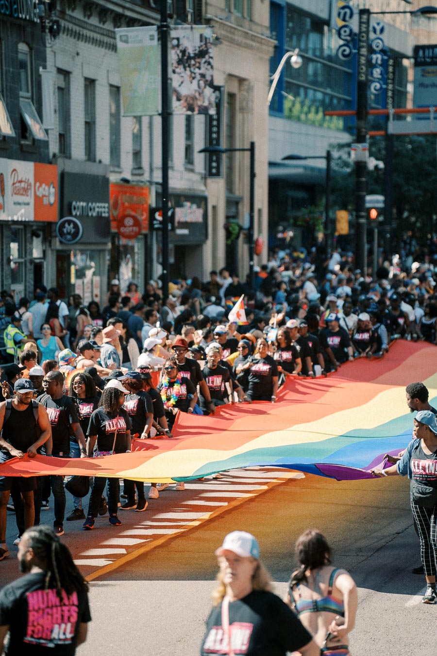 Participants in a pride parade march down a city street, carrying a large rainbow flag symbolizing LGBTQ+ pride and unity.