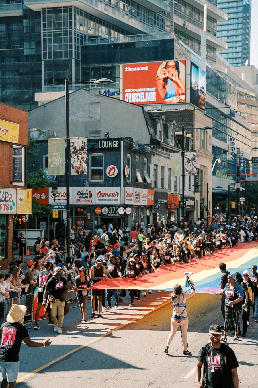Participants in a street parade carry a large rainbow flag, symbolizing LGBTQ+ pride, as spectators line the street. The event takes place in an urban setting with high-rise buildings and a digital billboard in the background.